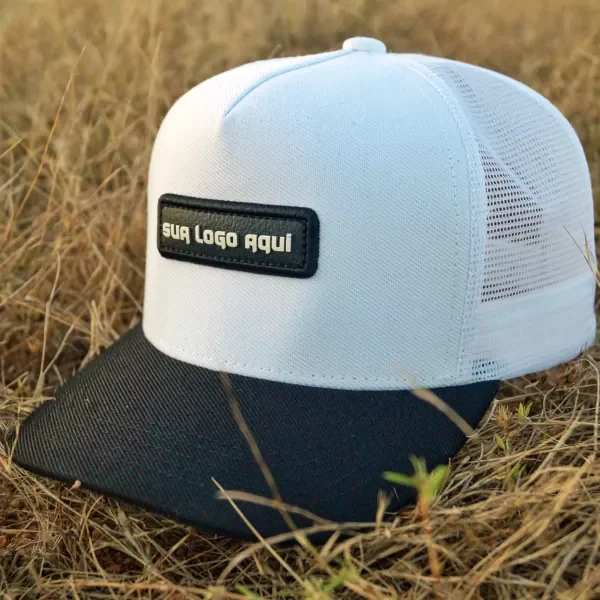 Boné trucker branco com aba preta e patch retangular para a sua logo, sobre capim seco