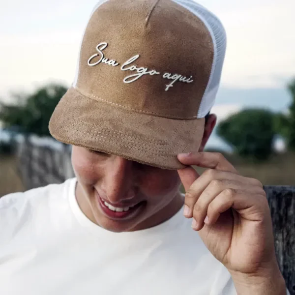 Jovem sorrindo segurando a aba de um boné trucker de camurça caramelo com logo bordada em branco