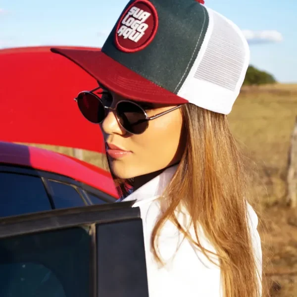 Mulher de óculos escuros usando boné trucker verde-escuro com aba vermelha e patch redondo para a sua logo