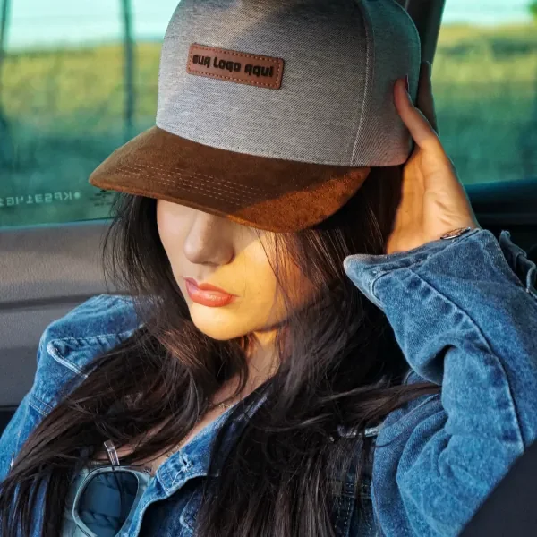 Mulher de jaqueta jeans segurando boné cinza com aba de camurça marrom e patch de couro
